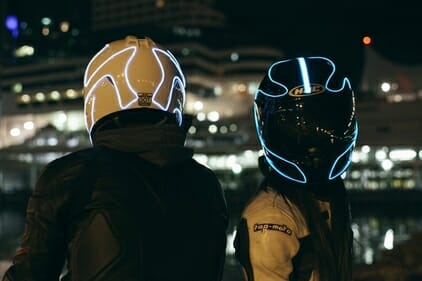 Two motorcyclists at nighttime, with reflective tape on their helmets to increase visiblity. 