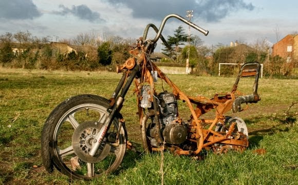 A close up of a rusty motorcycle, sitting in a field behind some houses.